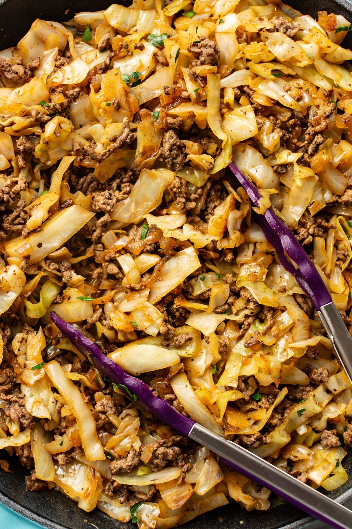 a skillet with ground beef and cabbage with kitchen tongs